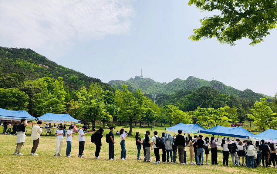 많은 학생이 버들골에서의 피크닉을 위해 돗자리를 빌리려 줄을 서 있다.